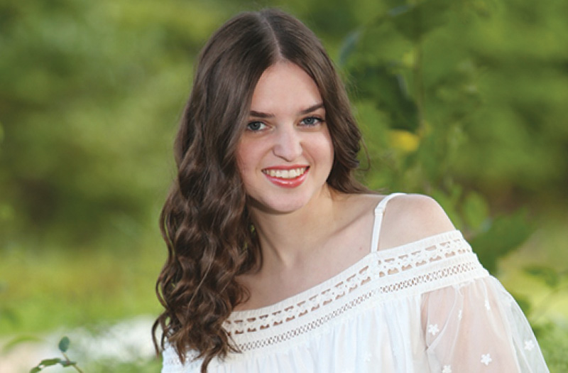 Marywood University student with curly brown hair in a white off-shoulder dress, standing against green foliage.